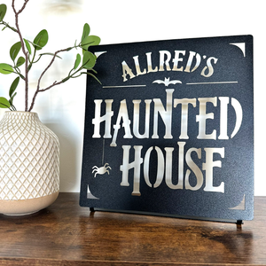A decorative metal sign reading \“Allred's Haunted House\“ features Halloween-themed elements like a bat and spider, displayed next to a textured vase with branches.
