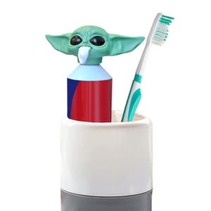 A toothpaste dispenser shaped like Baby Yoda's head dispenses toothpaste through its mouth, sitting in a holder alongside a teal and white toothbrush.