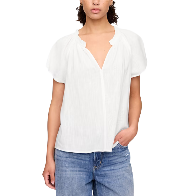 A woman wears a white short-sleeve blouse with a scalloped V-neck and relaxed fit, paired with blue jeans.