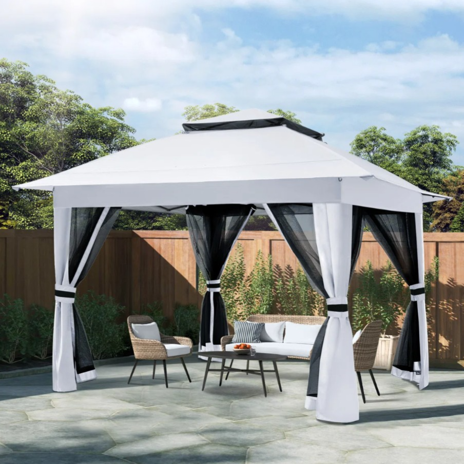 A white outdoor gazebo with black mesh curtains is set up on a paved area, accompanied by wicker chairs and a coffee table, surrounded by a wooden fence and greenery.