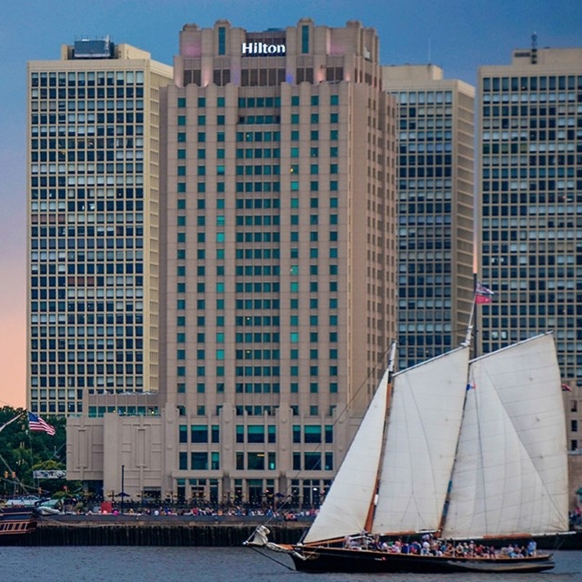 A large Hilton hotel building with adjacent skyscrapers, in front of which a sailboat is navigating on the water.
