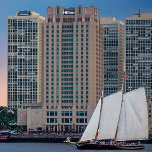 A large Hilton hotel building with adjacent skyscrapers, in front of which a sailboat is navigating on the water.