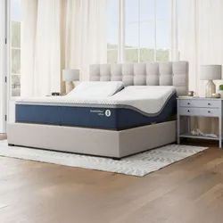 Adjustable bed with a tufted headboard, ComfortNext Ultra mattress, and two matching side tables, set in a bright room with wooden flooring and white curtains.