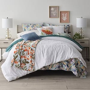 The 3pc Crinkle Comforter Set features a textured white comforter with a floral printed reverse, two matching shams, and coordinating floral and solid decorative pillows. Accompanied by a floral throw, the bedding is styled with neutral side tables and lamps.