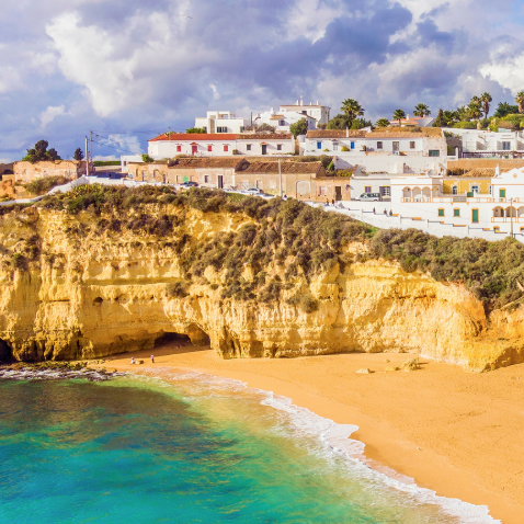 A picturesque coastal scene featuring golden cliffs, a sandy beach, and turquoise water, with white buildings perched atop the cliffs under a partly cloudy sky.