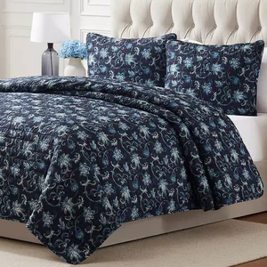 A bed features a blue floral-patterned quilt with matching pillow shams, set against a tufted headboard. A lamp and a white vase with blue flowers are on a side table.