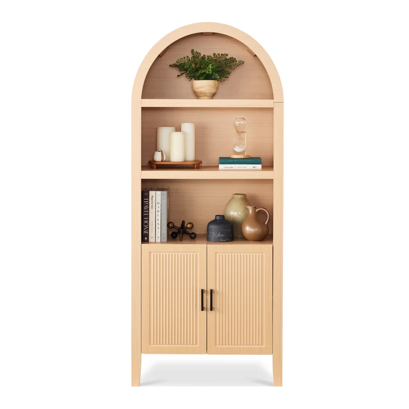A tall, arched wooden bookcase with shelves containing candles, a plant, an hourglass, books, and ceramic vases. The lower section features two textured cabinet doors with metal handles.