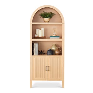 A tall, arched wooden bookcase with shelves containing candles, a plant, an hourglass, books, and ceramic vases. The lower section features two textured cabinet doors with metal handles.