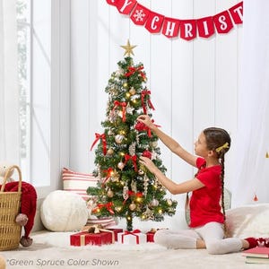 A 4-foot pre-lit Christmas tree is adorned with red and gold ornaments, a gold star topper, and surrounded by gifts. A girl is decorating the tree.