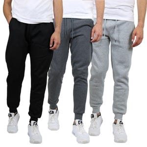 A set of three men's fleece joggers in black, dark grey, and light grey. They feature drawstrings, elastic waistbands, and tapered cuffs, styled with white sneakers and white t-shirts.