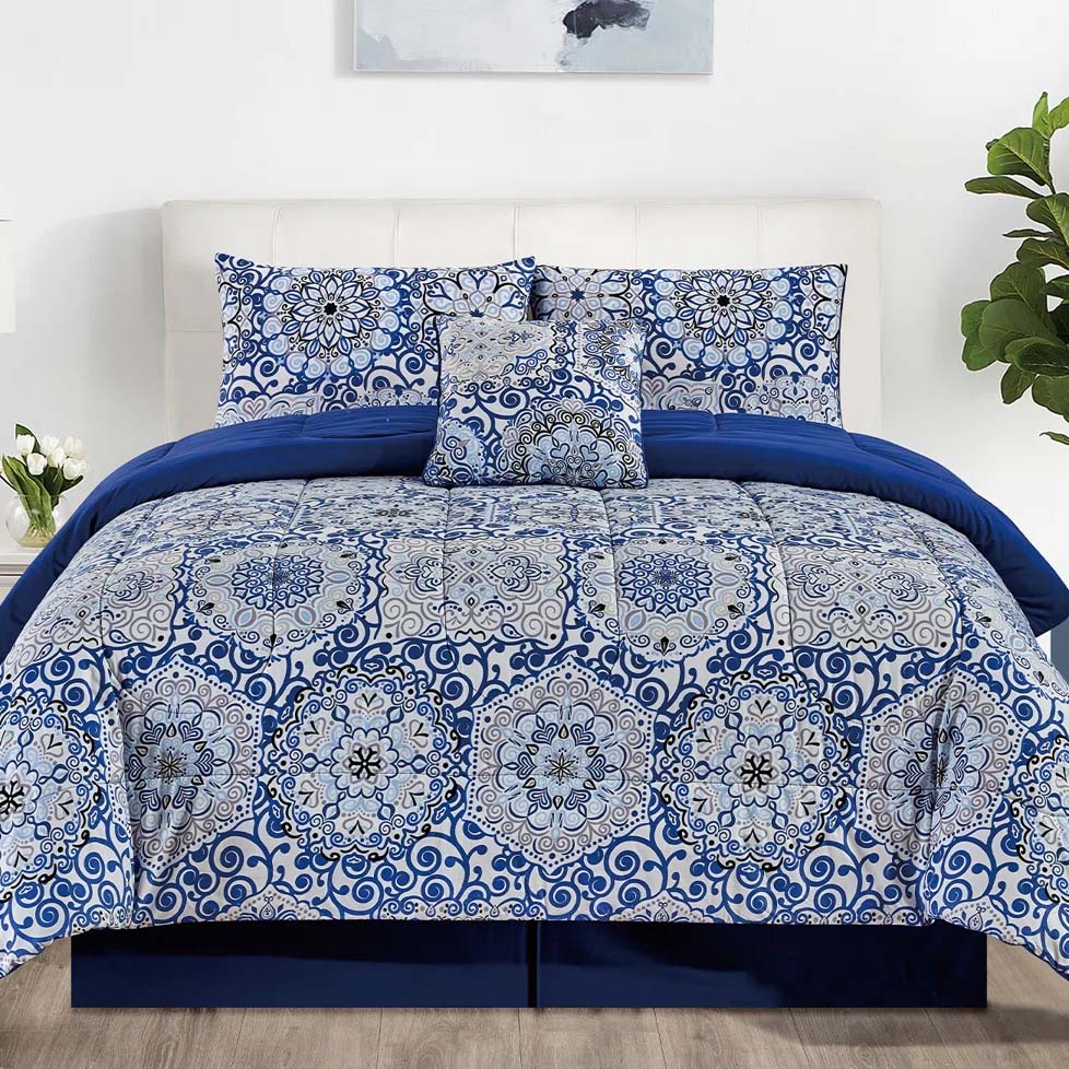 Blue and white bedding set with intricate mandala patterns, including a comforter, pillowcases, and decorative pillow. Room features white walls, wood floor, and plants.