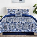 Blue and white bedding set with intricate mandala patterns, including a comforter, pillowcases, and decorative pillow. Room features white walls, wood floor, and plants.