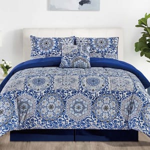 Blue and white bedding set with intricate mandala patterns, including a comforter, pillowcases, and decorative pillow. Room features white walls, wood floor, and plants.