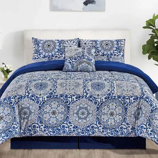 Blue and white bedding set with intricate mandala patterns, including a comforter, pillowcases, and decorative pillow. Room features white walls, wood floor, and plants.