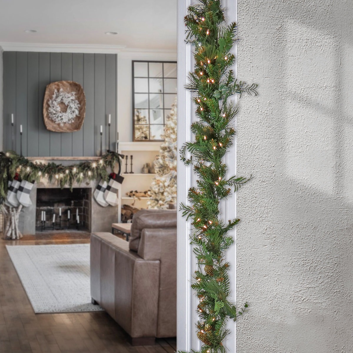 A holiday-themed living room features a fireplace with garland, stockings, and a wreath. A doorway is adorned with a lighted garland.