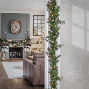 A holiday-themed living room features a fireplace with garland, stockings, and a wreath. A doorway is adorned with a lighted garland.