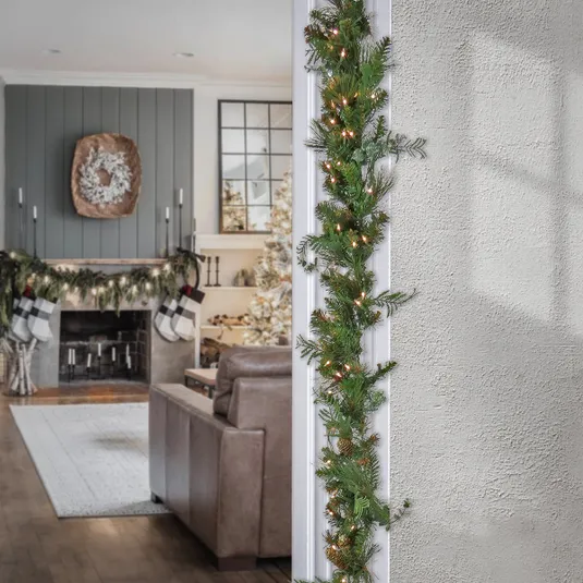A holiday-themed living room features a fireplace with garland, stockings, and a wreath. A doorway is adorned with a lighted garland.