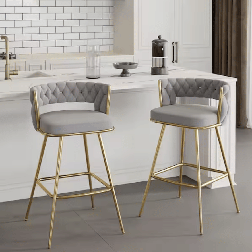 Two bar stools with gray cushioned seats and tufted backs, featuring gold metal frames, are positioned in a modern kitchen setting.