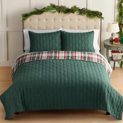 A bed with a green quilted comforter set, featuring matching pillowcases and a plaid accent throw. Greenery decorates the headboard, and a small side table holds holiday-themed items.