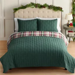 A bed with a green quilted comforter set, featuring matching pillowcases and a plaid accent throw. Greenery decorates the headboard, and a small side table holds holiday-themed items.