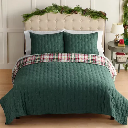 A bed with a green quilted comforter set, featuring matching pillowcases and a plaid accent throw. Greenery decorates the headboard, and a small side table holds holiday-themed items.
