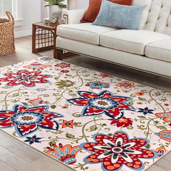 A living room area features a cream-colored sofa with a decorative rug on the floor, displaying vibrant floral patterns in red, blue, and orange hues. A small wooden coffee table and a basket are also visible in the bright, sunlit space.