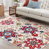 A living room area features a cream-colored sofa with a decorative rug on the floor, displaying vibrant floral patterns in red, blue, and orange hues. A small wooden coffee table and a basket are also visible in the bright, sunlit space.