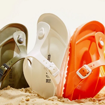 Three pairs of Birkenstock Gizeh EVA sandals in olive, white, and orange are displayed on a sandy surface, each featuring a single strap and a thong design.