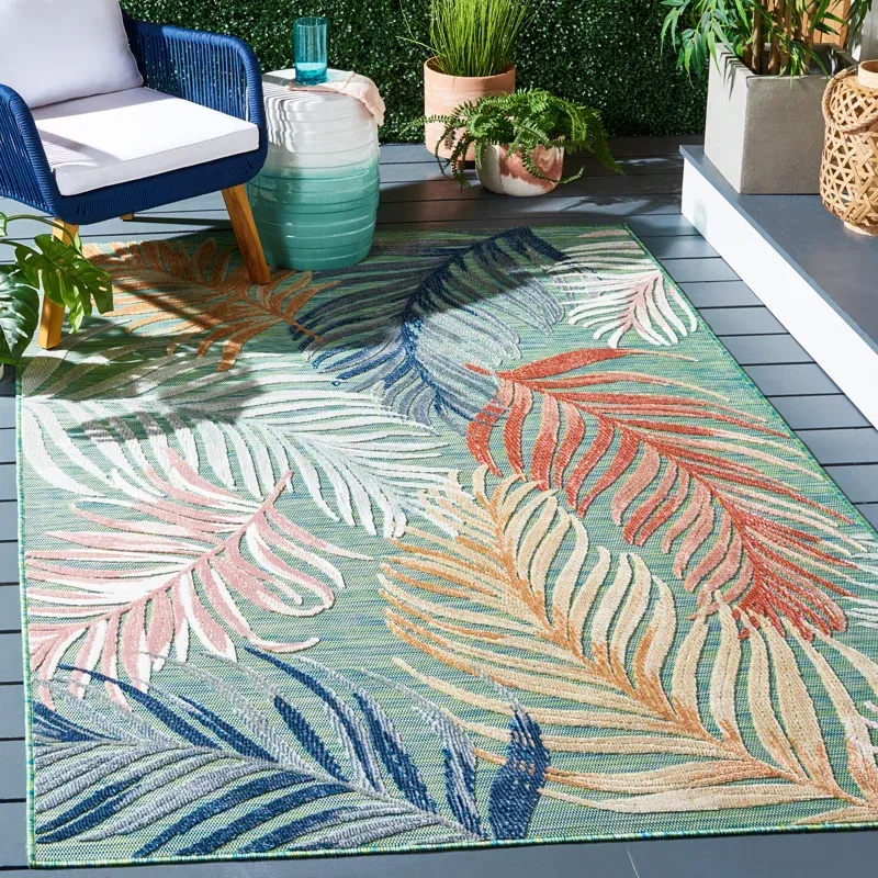 A colorful outdoor rug with a tropical leaf pattern is shown on a patio, accompanied by a blue-and-white chair, a stack of layered glass-like tables, and various plants in pots.