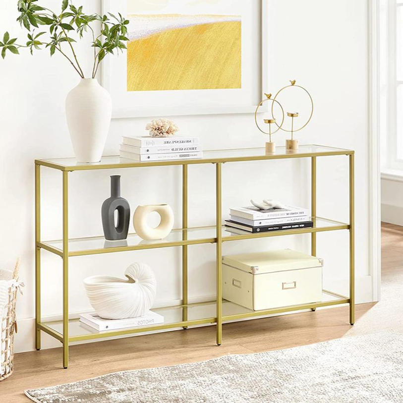 A gold metal console table with two glass shelves displays decorative items, including vases, books, and a box. The setting features a beige rug, a plant in a basket, and a framed art piece on the wall.