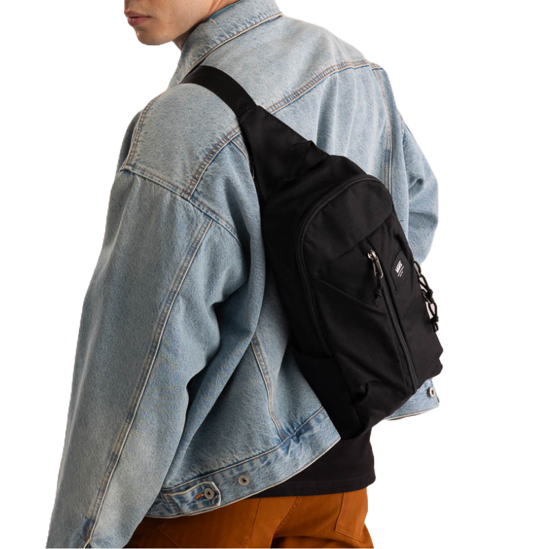 A black sling bag with a prominent white Vans logo, featuring multiple zip compartments, worn over the shoulder and across the back.