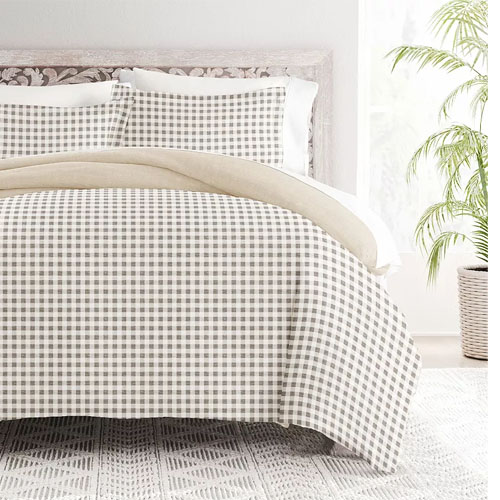 A checkered bedding set with a beige and white pattern, including a duvet cover and pillowcases, is styled in a bright room with a potted plant and a patterned rug.