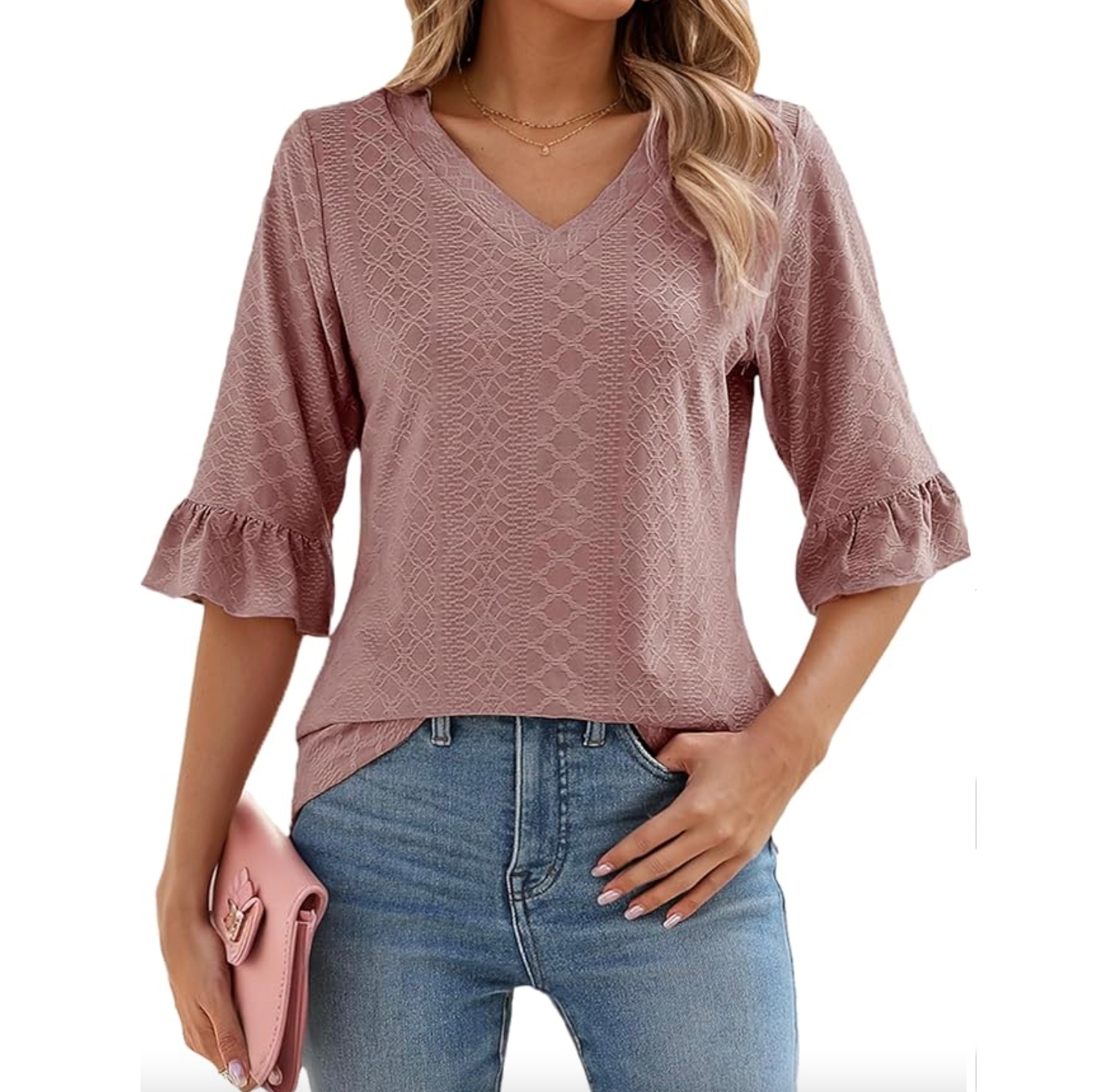 A woman is wearing a textured, dusty rose V-neck blouse with three-quarter sleeves and ruffle details, paired with blue jeans. She's holding a pink clutch and wearing a delicate necklace.