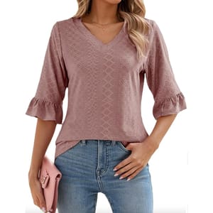 A woman is wearing a textured, dusty rose V-neck blouse with three-quarter sleeves and ruffle details, paired with blue jeans. She's holding a pink clutch and wearing a delicate necklace.