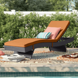 Poolside lounge chair with a brown cushion and two pillows, one striped and one solid. It sits on a patterned outdoor rug beside a pool, surrounded by greenery.