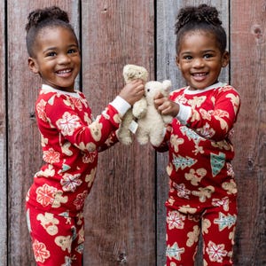 Two young children wear red pajamas with holiday-themed designs, including gingerbread men and Christmas trees, while holding plush teddy bears in front of a wooden backdrop.
