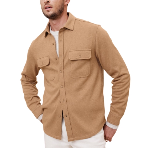 Tan twill shirt jacket featuring a button-up front, two chest pockets with flaps, and long sleeves. Worn over a white shirt, paired with white pants.