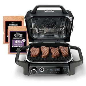 Ninja Woodfire Grill with integrated control panel, featuring four grilled steaks on the cooking surface and accompanied by Ninja Woodfire Pellets in two flavor blends.