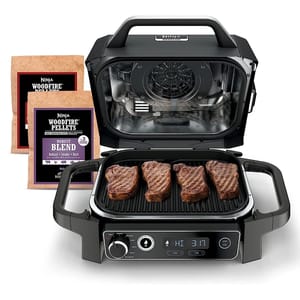 Ninja Woodfire Grill with integrated control panel, featuring four grilled steaks on the cooking surface and accompanied by Ninja Woodfire Pellets in two flavor blends.