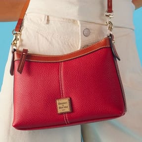 A red Dooney & Bourke crossbody bag with brown leather trim and gold hardware, worn over white pants.