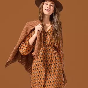 A woman is wearing a long sleeve dress with a diamond-shaped pattern in brown tones, paired with a matching fringed shawl and a wide-brimmed hat.