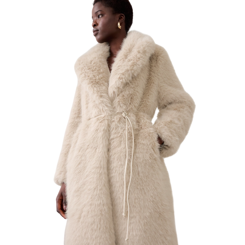 A woman wears a beige, fluffy faux fur coat with long sleeves and a soft, plush texture. The coat features a wrap style with a waist tie and a large collar.