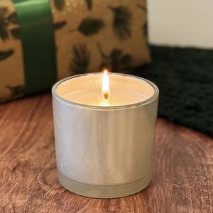 A lit candle in a silver container sits on a wooden surface. A wrapped gift is in the background.