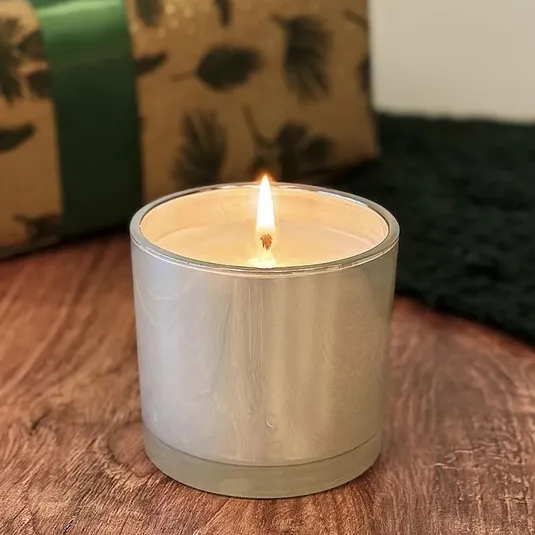 A lit candle in a silver container sits on a wooden surface. A wrapped gift is in the background.