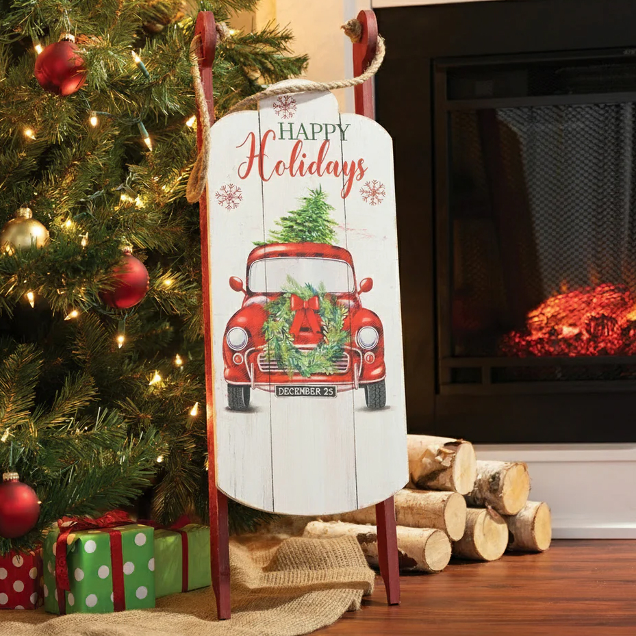 Festive holiday decoration featuring a wooden sled with a vintage red car design, adorned with a wreath and Christmas tree, placed beside a Christmas tree and fireplace.