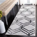 A white runner rug with a black geometric pattern is placed on a dark wooden floor, next to a beige tufted bench with chrome legs. A white vase with yellow flowers is visible in the foreground.
