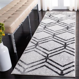 A white runner rug with a black geometric pattern is placed on a dark wooden floor, next to a beige tufted bench with chrome legs. A white vase with yellow flowers is visible in the foreground.