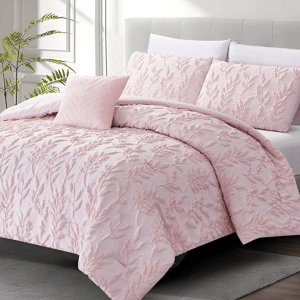 A light pink comforter set with a textured floral pattern, featuring one comforter, two standard shams, and a small accent pillow, displayed on a neatly made bed with a neutral headboard and a plant in the background.
