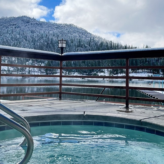 A hot tub sits on a patio overlooking a snowy mountain landscape, with steam rising from the warm water.