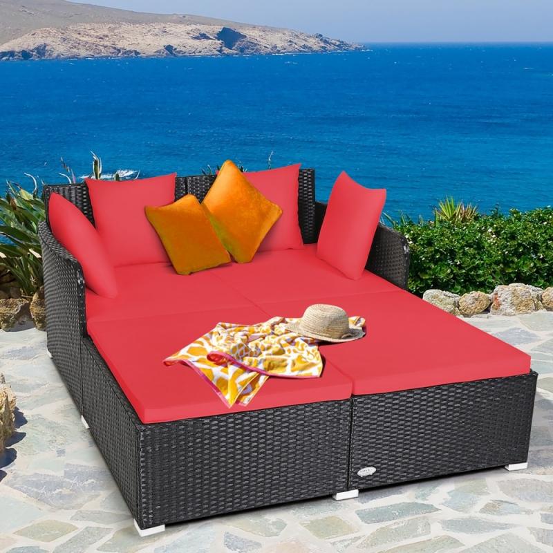 A stylish outdoor patio daybed with a black wicker frame and vibrant red cushions, featuring a relaxing layout adorned with assorted pillows, a sunhat, and a towel, set against a scenic ocean backdrop.